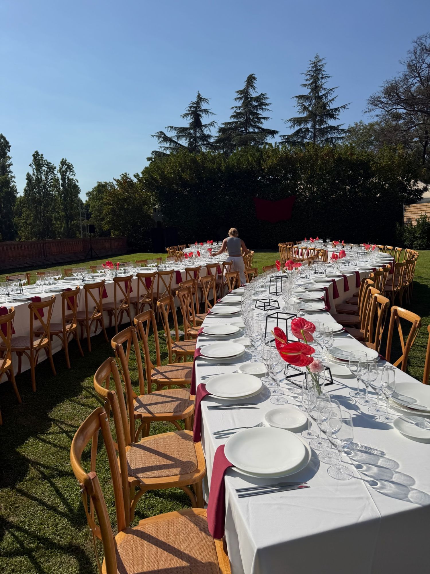 Long outdoor banquet tables in white linen with burgundy napkins and red anthuriums, set on the lawn